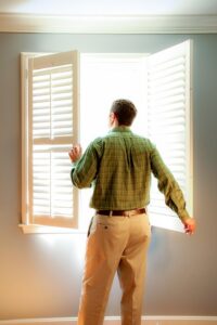 Man facing away standing in front of an open window, looking out and holding the shutters open.